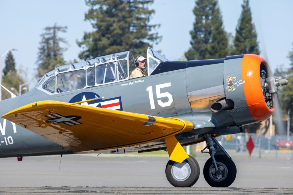 Classic World War II era aircraft on runway with pilot, clear blue sky and trees in background.