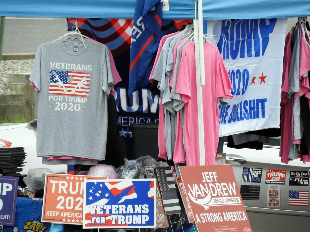 A stall displaying Trump 2020 merchandise including shirts and signs at an outdoor market.