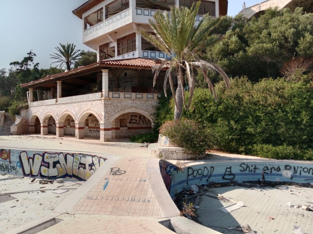 Deserted villa with graffiti and empty pool, surrounded by greenery under a clear sky.